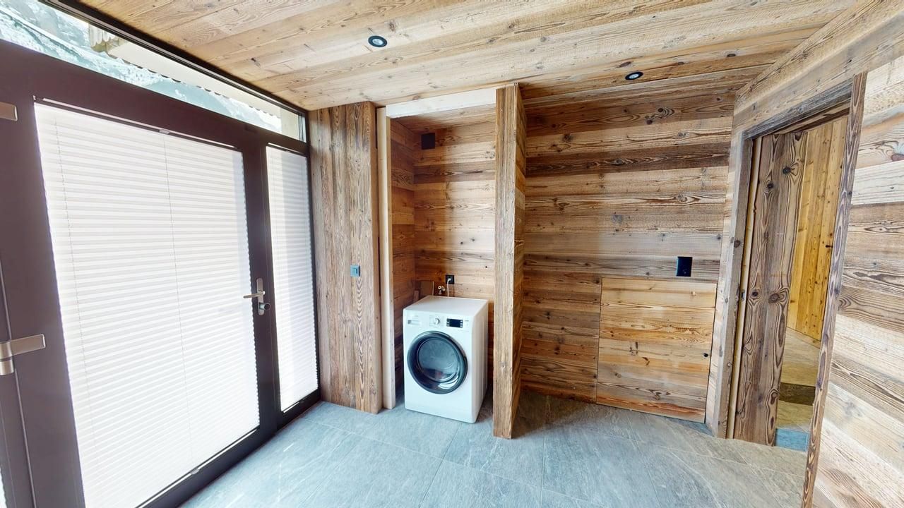 A modern laundry area with a white washing machine set against rustic light-wood paneled walls and a tiled floor.