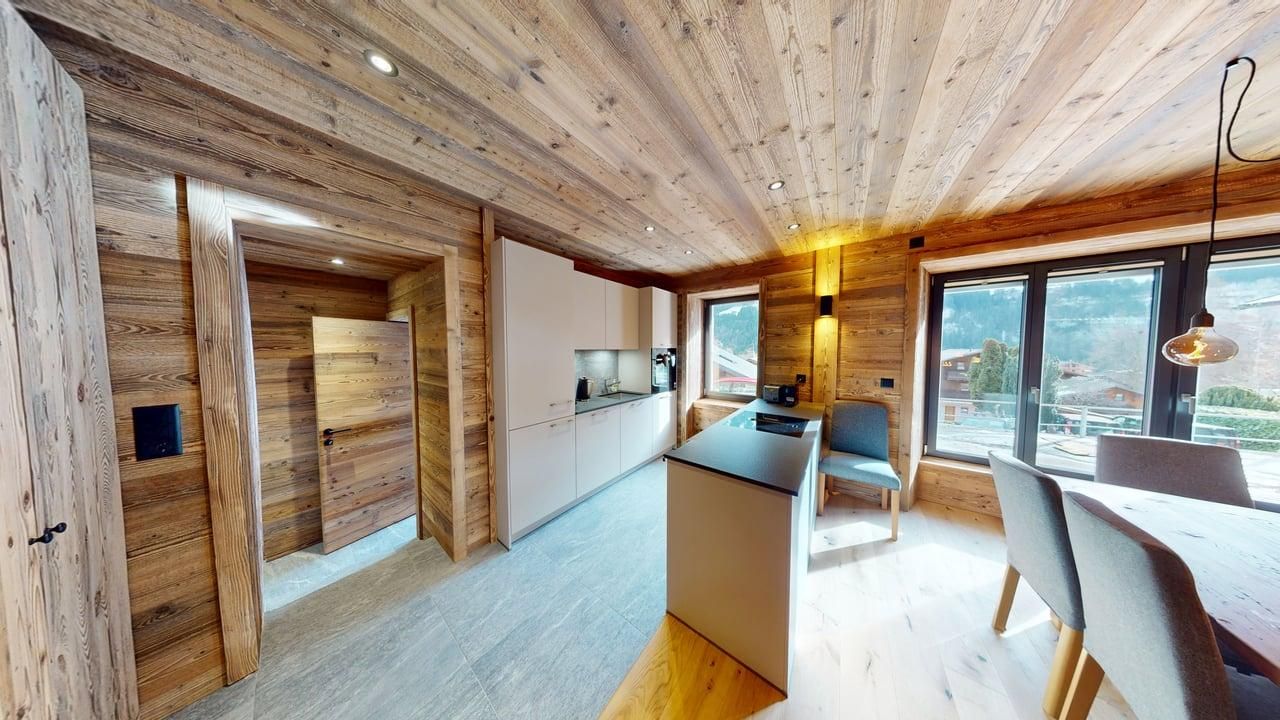 Une cuisine moderne et lumineuse dans un chalet rustique aux murs lambrissés, avec un îlot central, un coin repas et de grandes fenêtres.