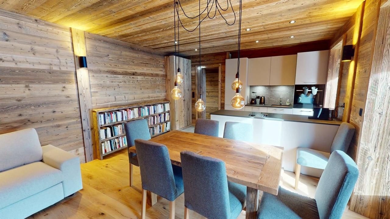 Un salon-salle à manger rustique avec des murs lambrissés, une table à manger avec des chaises bleues et une cuisine ouverte.