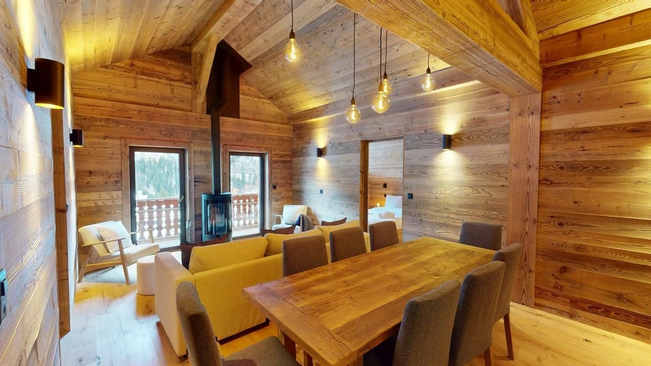 A rustic, wooden interior featuring a dining table with chairs, a seating area with a fireplace, and warm ambient lighting.