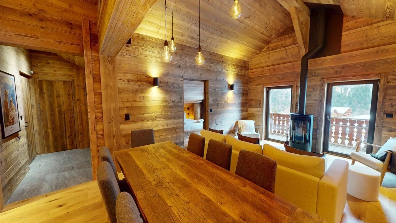 A rustic, wood-paneled living and dining room with a long wooden table, a light-colored sofa, and a tall wood-burning stove.