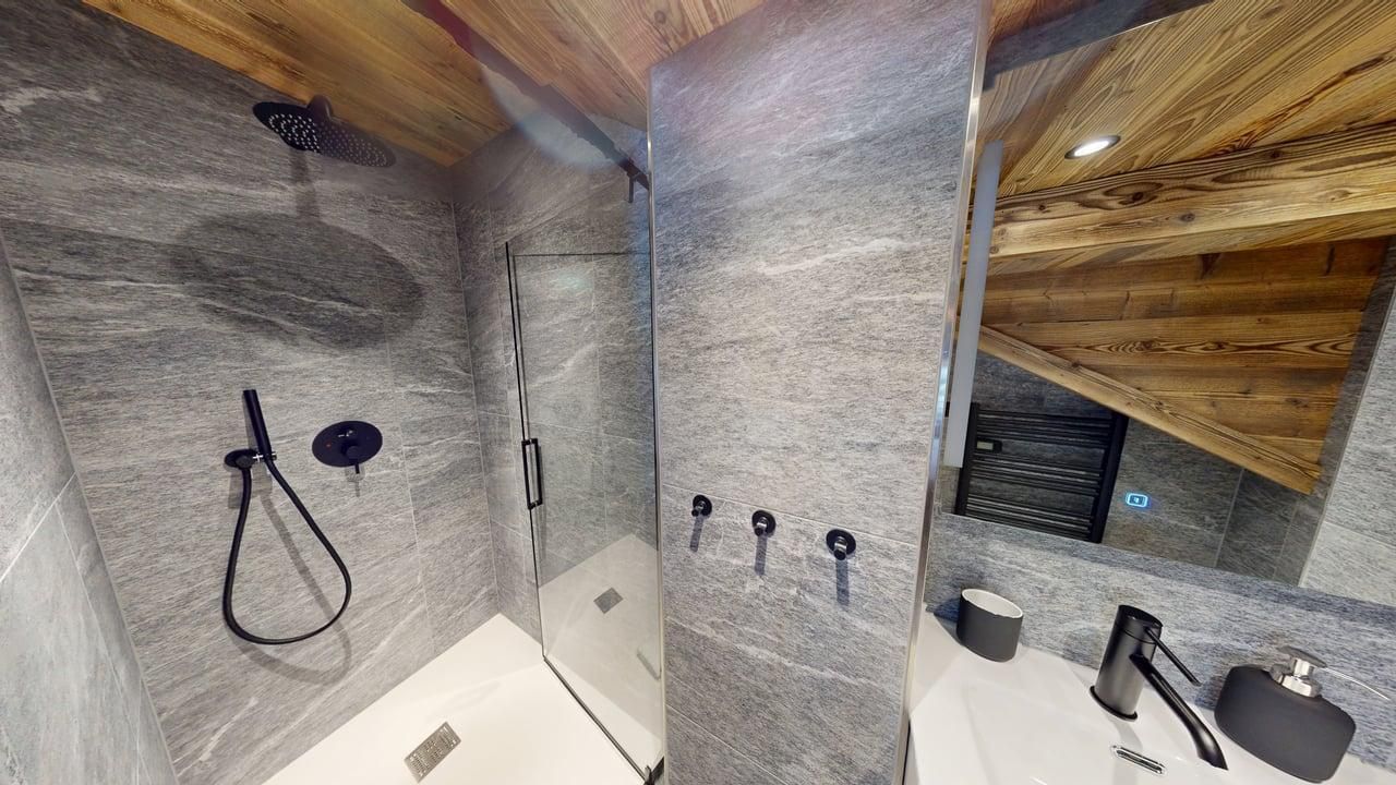 A modern bathroom with grey stone walls, a glass-enclosed shower, black fixtures, and a wood-paneled slanted ceiling.