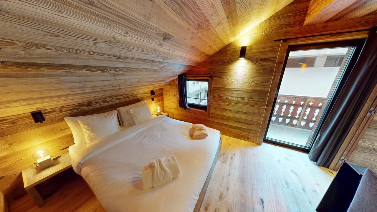 A cozy bedroom with wooden walls and ceiling, featuring a bed with white linens and a balcony door overlooking a snowy area.