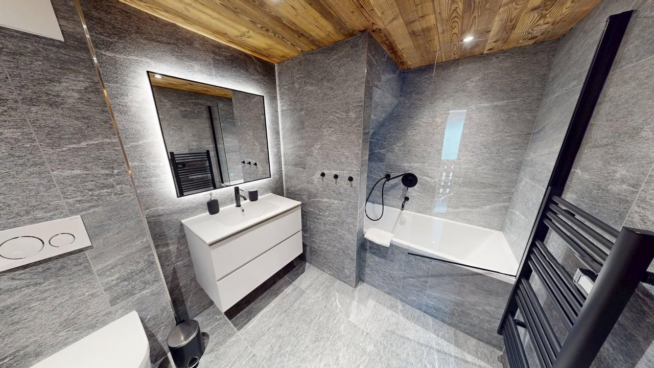 Modern bathroom with gray tiled walls, a white vanity with an illuminated mirror, a bathtub, and a black towel warmer.