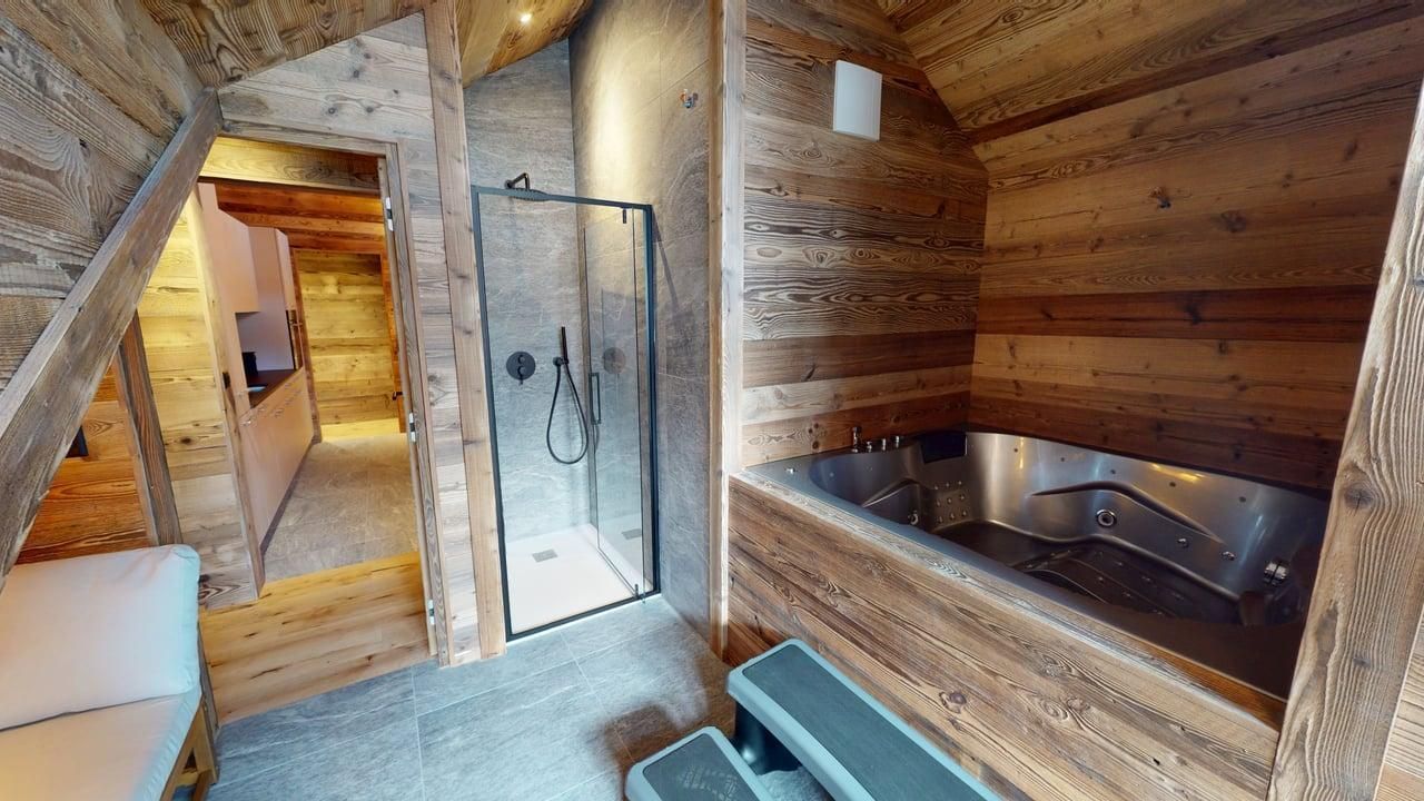 A rustic wooden bathroom with a built-in spa tub, a glass-enclosed shower, and steps leading to a connecting hallway.