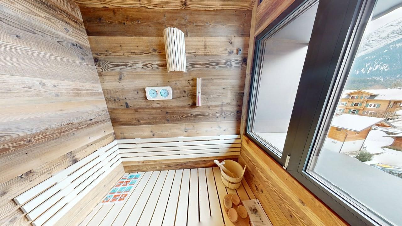 A wooden sauna interior with a slat floor, a wall-mounted bucket, and a large window looking out onto a snowy landscape.