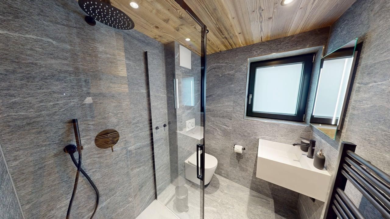 Modern grey stone bathroom with a walk-in shower, wall-mounted toilet, vanity sink, and a wooden paneled ceiling.