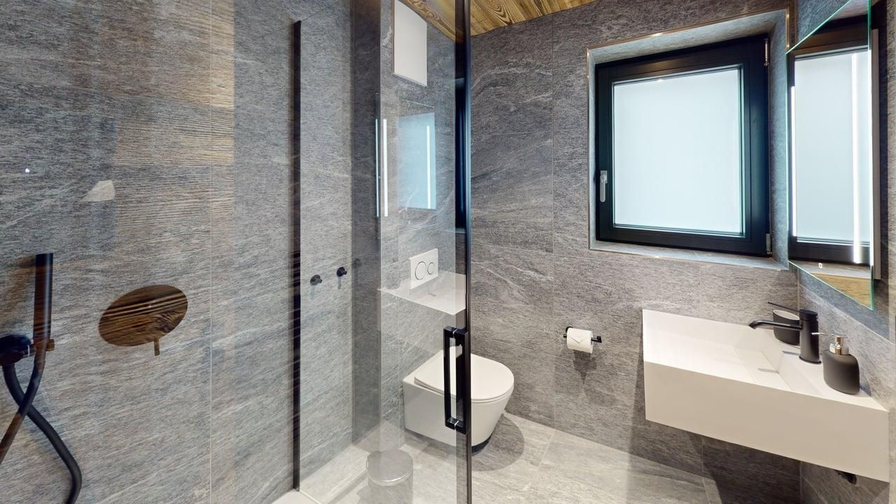Modern bathroom with grey stone walls, a glass-enclosed shower, a white wall-mounted sink, and a toilet.