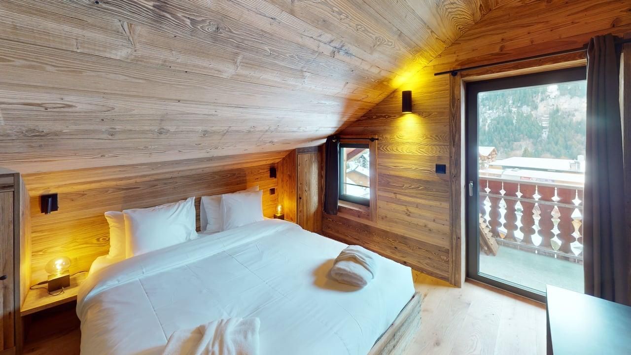 A cozy attic bedroom with wood-paneled walls, a white-made bed, warm lighting, and a glass door opening to a snowy balcony.