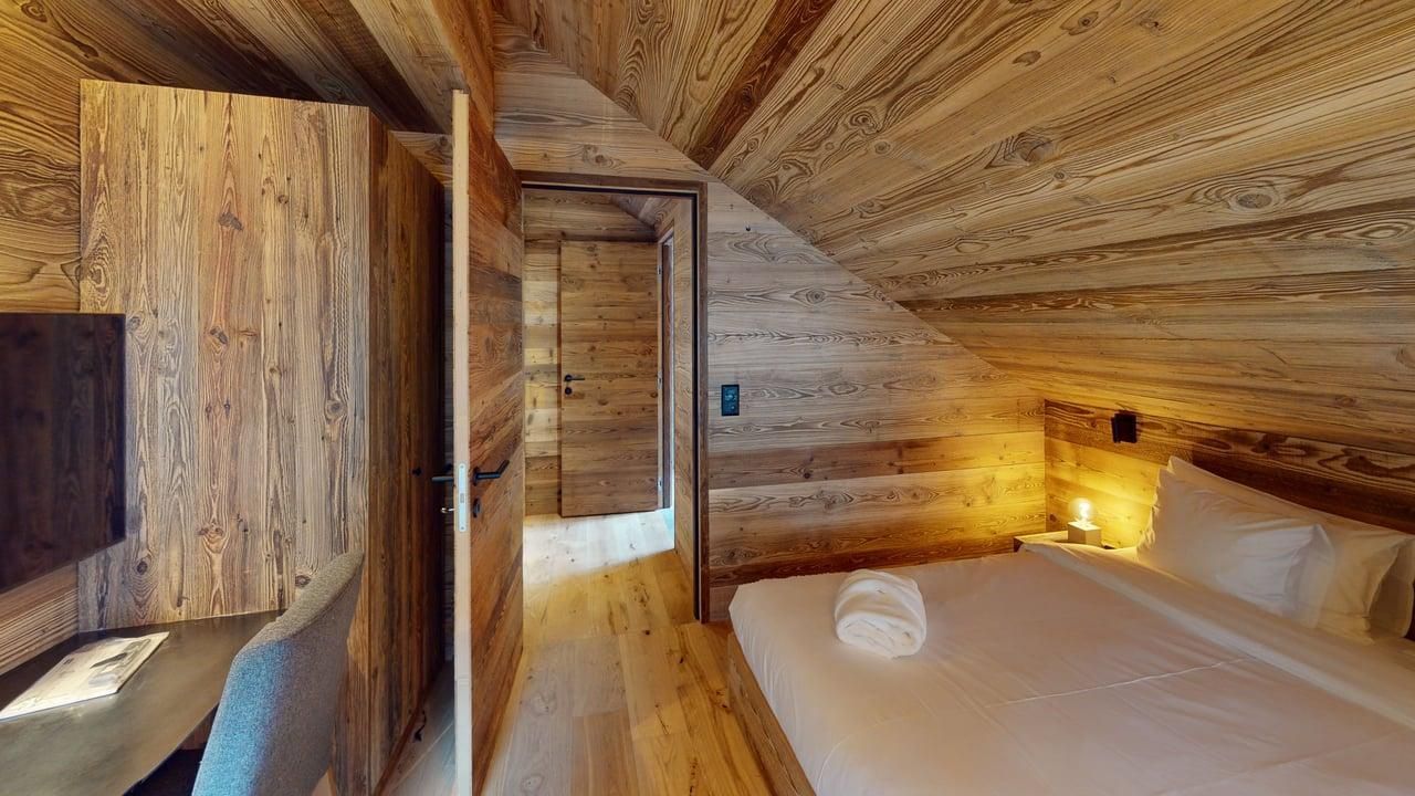 A cozy bedroom with wooden walls and ceiling, featuring a bed with white linens and a small desk in a rustic attic room.