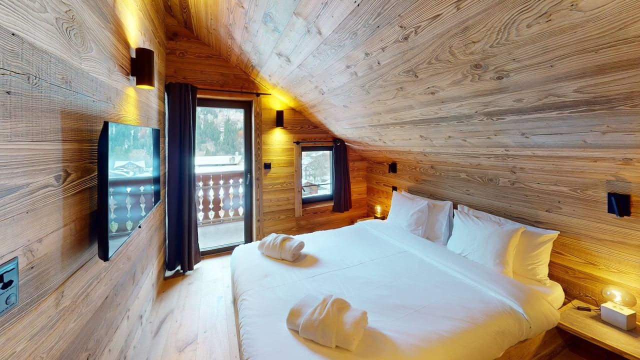 A cozy bedroom with wooden walls and ceiling, featuring a bed with white linens, a TV, and a balcony door.