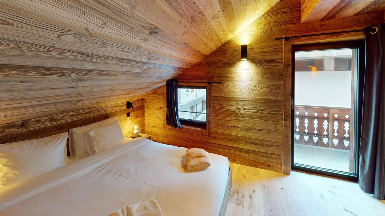 A cozy attic bedroom with a white bed, wood-paneled walls, a small window, and a glass door opening to a balcony.