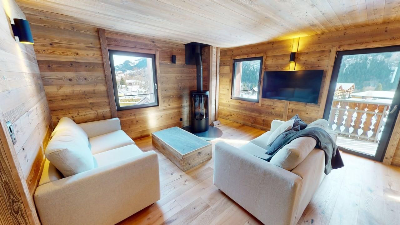 Salon rustique et chaleureux de chalet avec canapés clairs, murs lambrissés, cheminée et vue depuis un balcon.