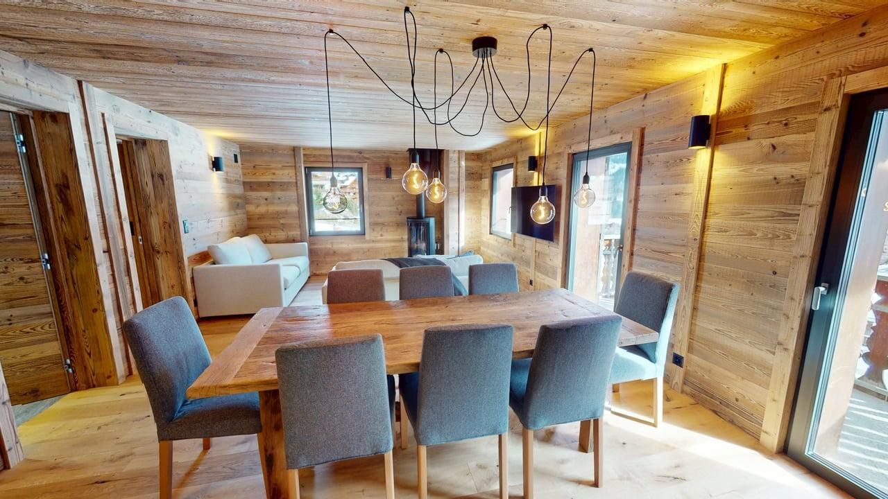 Une salle à manger avec une table en bois, six chaises grises et un lustre araignée unique, dans un intérieur rustique de style cabane en rondins.