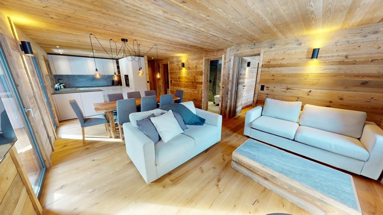 Un espace de vie avec des murs et des plafonds lambrissés, comprenant deux canapés gris clair, une table à manger en bois et une cuisine.