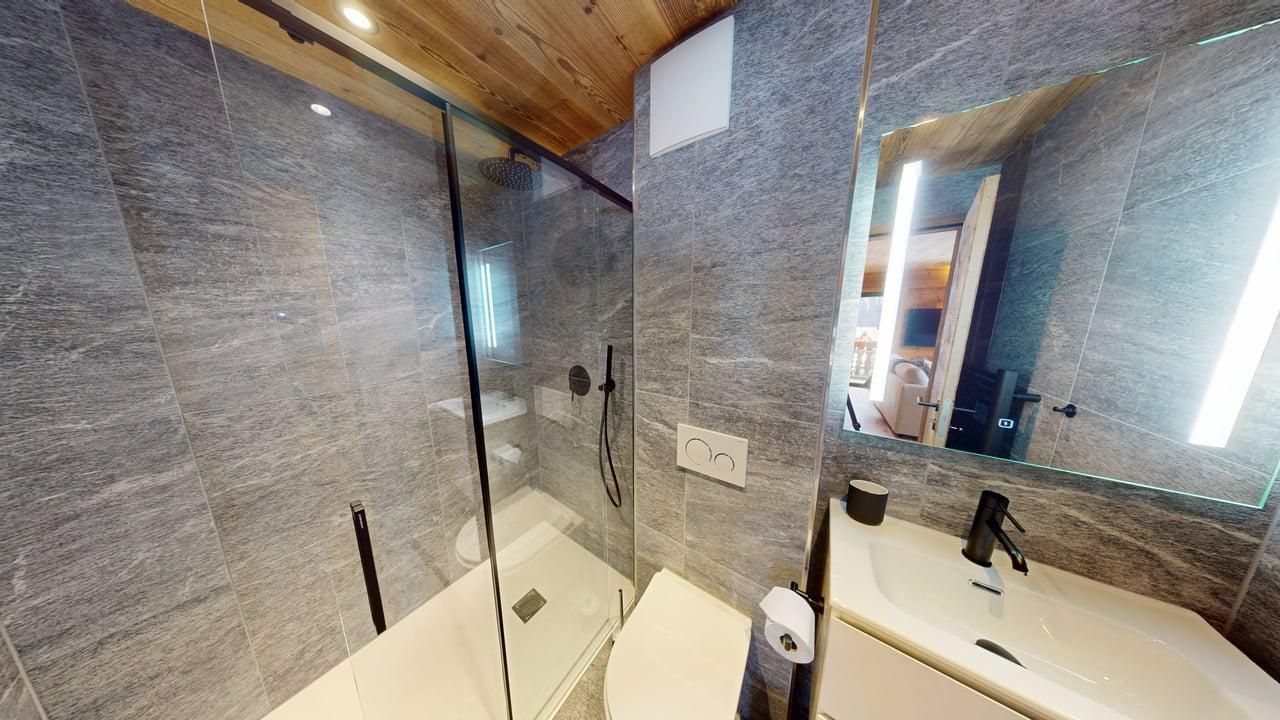 Une salle de bains moderne avec une douche vitrée, des murs carrelés gris, un lavabo blanc, des toilettes et un plafond en bois.