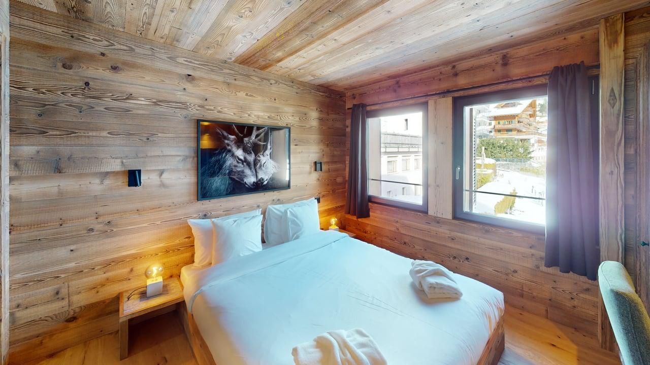 Une chambre confortable avec des murs et un plafond en bois, un lit blanc, des tableaux et des fenêtres donnant sur un paysage enneigé.