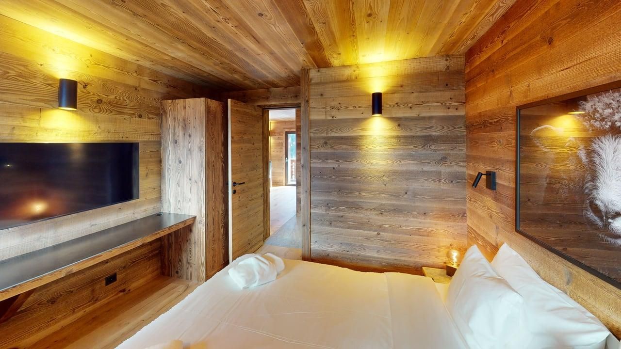 Une chambre rustique avec des murs lambrissés, un grand lit aux draps blancs, une télévision fixée au mur et un éclairage d'ambiance chaleureux.