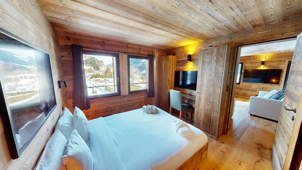 Une chambre rustique aux murs lambrissés, avec un grand lit, des fenêtres donnant sur un paysage enneigé et un salon attenant.