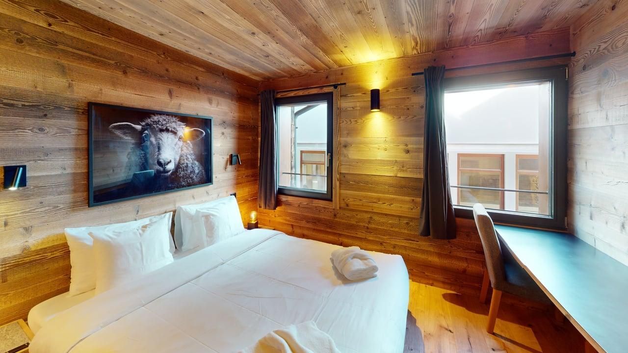 Une chambre confortable avec des murs en bois, un lit aux draps blancs, une estampe de mouton encadrée et un bureau avec une chaise près de la fenêtre.