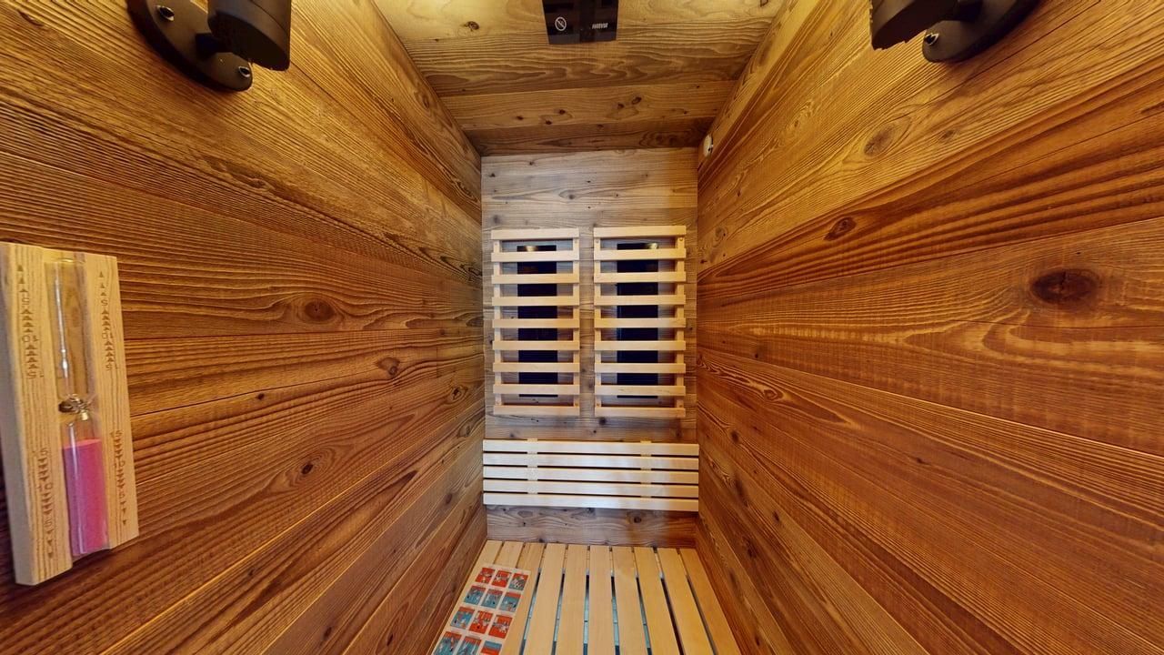 A small sauna interior featuring light-toned wooden paneling, a wooden bench, slatted walls, and an hourglass on the wall.