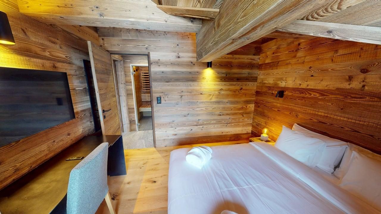 A rustic bedroom with wooden walls and ceiling, featuring a bed with white linens, a desk, and an open doorway.