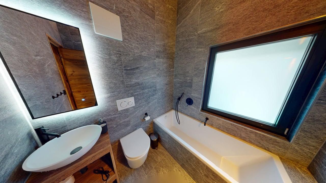 A modern bathroom featuring grey tiled walls, an oval vessel sink, a white toilet, and a bathtub under a frosted window.