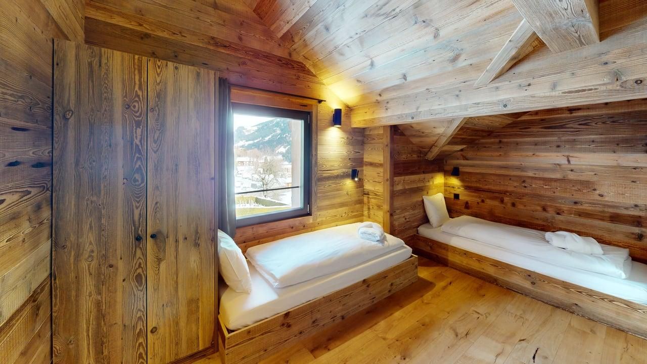 A rustic attic bedroom with wooden walls, ceiling beams, and two single beds set against a window and side wall.
