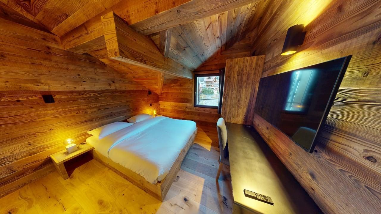 A cozy bedroom with wooden walls and ceiling, featuring a made bed, a small bedside lamp, a desk, and a wall-mounted TV.