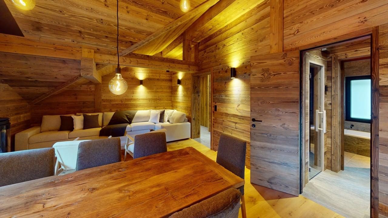 A rustic dining and living area featuring wood-paneled walls, a wooden table with chairs, and a sectional sofa in the back.