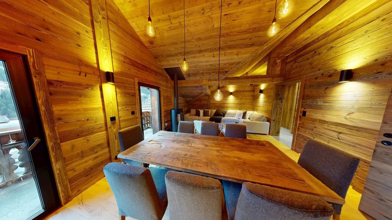 A rustic dining room with a large wooden table, gray upholstered chairs, and warm ambient lighting in a wood-paneled cabin.