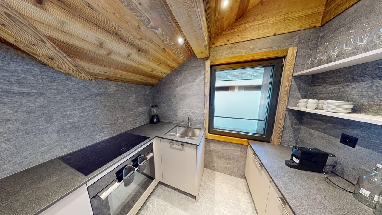 Modern kitchen with wood-paneled sloped ceiling, grey stone-like walls, white cabinets, and a sink by a window.
