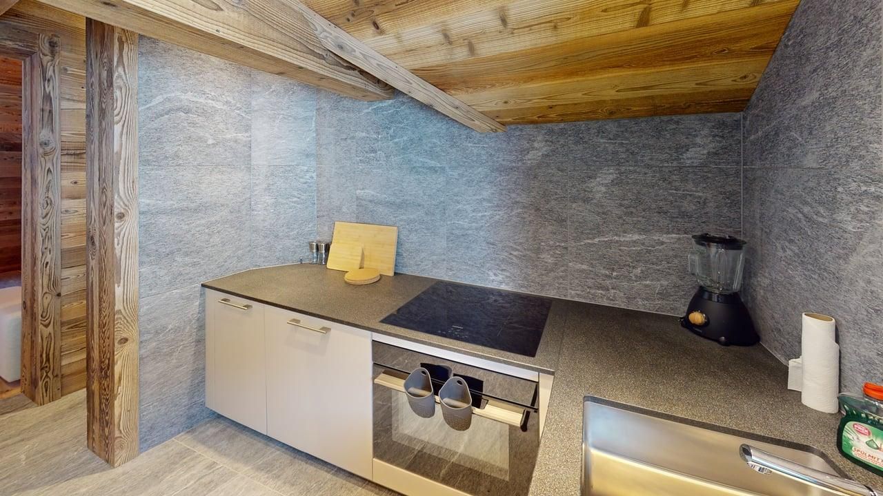 A modern kitchenette with stone walls, light cabinets, a black stovetop, an oven, and a sink beneath a wood-beamed ceiling.
