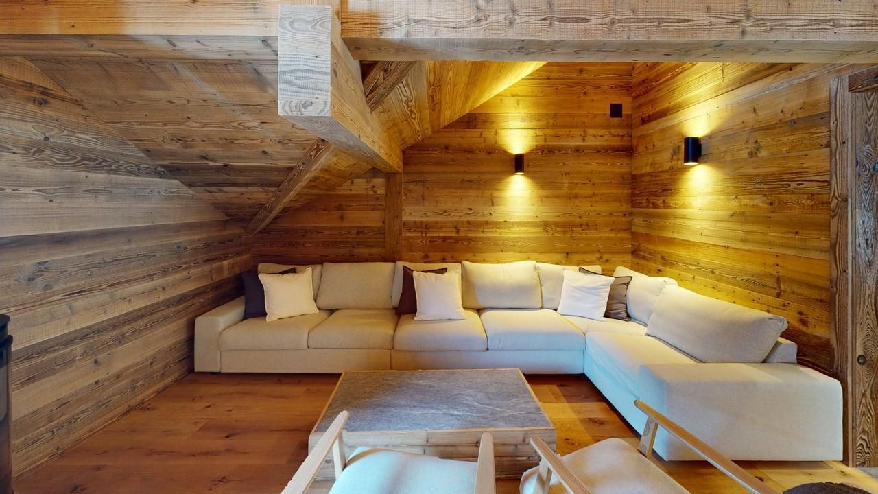 A cozy, rustic attic living room featuring wooden walls, a large white sectional sofa, and warm ambient wall lighting.
