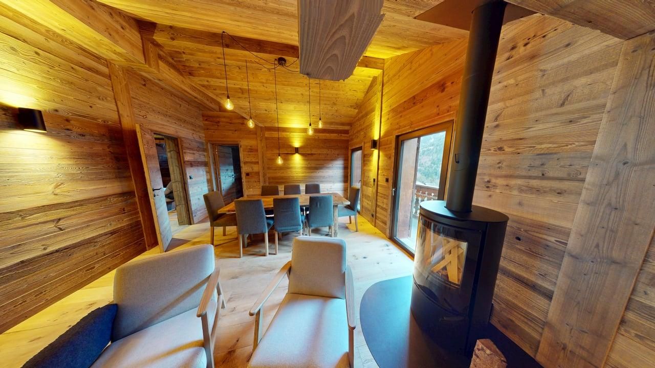 A rustic wood-paneled room features a dining area with chairs and a black wood-burning stove in the foreground.