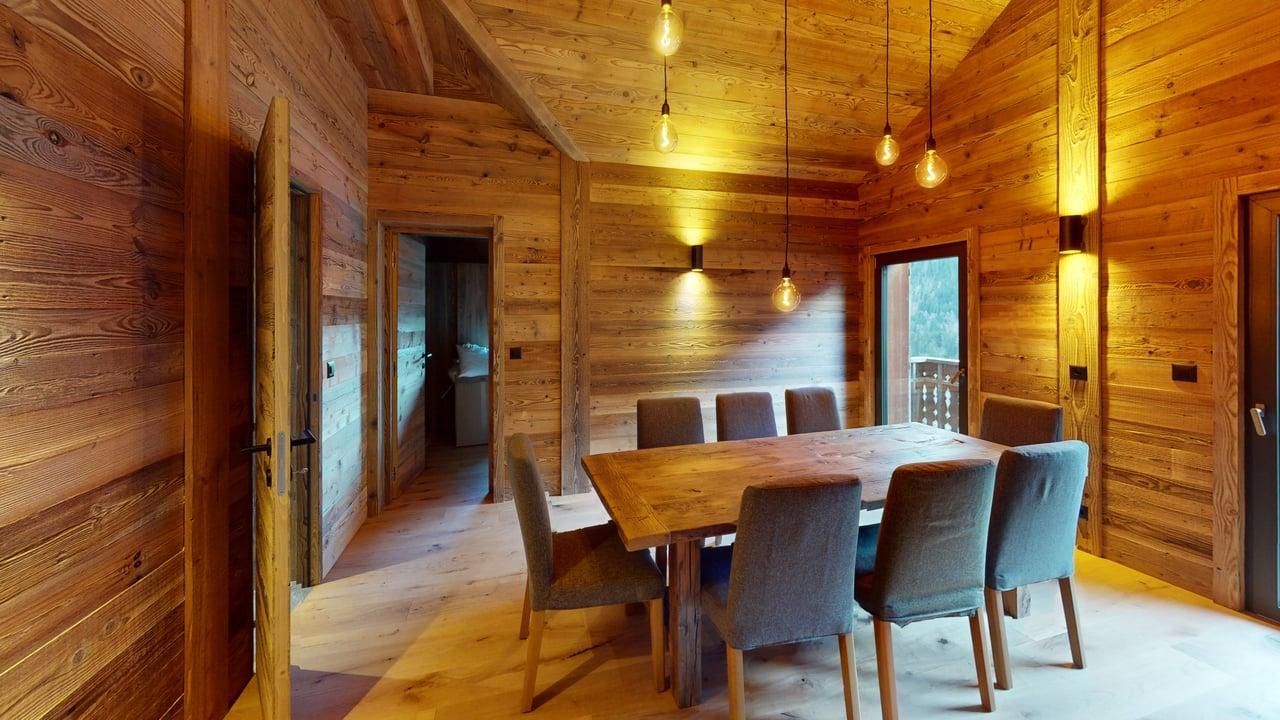 A rustic dining room with wooden walls and floors featuring a large table and six grey chairs under hanging pendant lights.