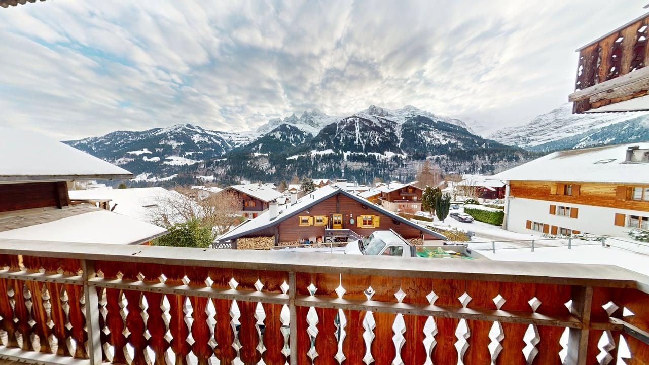 Une balustrade en bois surplombe un village de montagne enneigé, avec au loin de hauts sommets escarpés saupoudrés de neige.