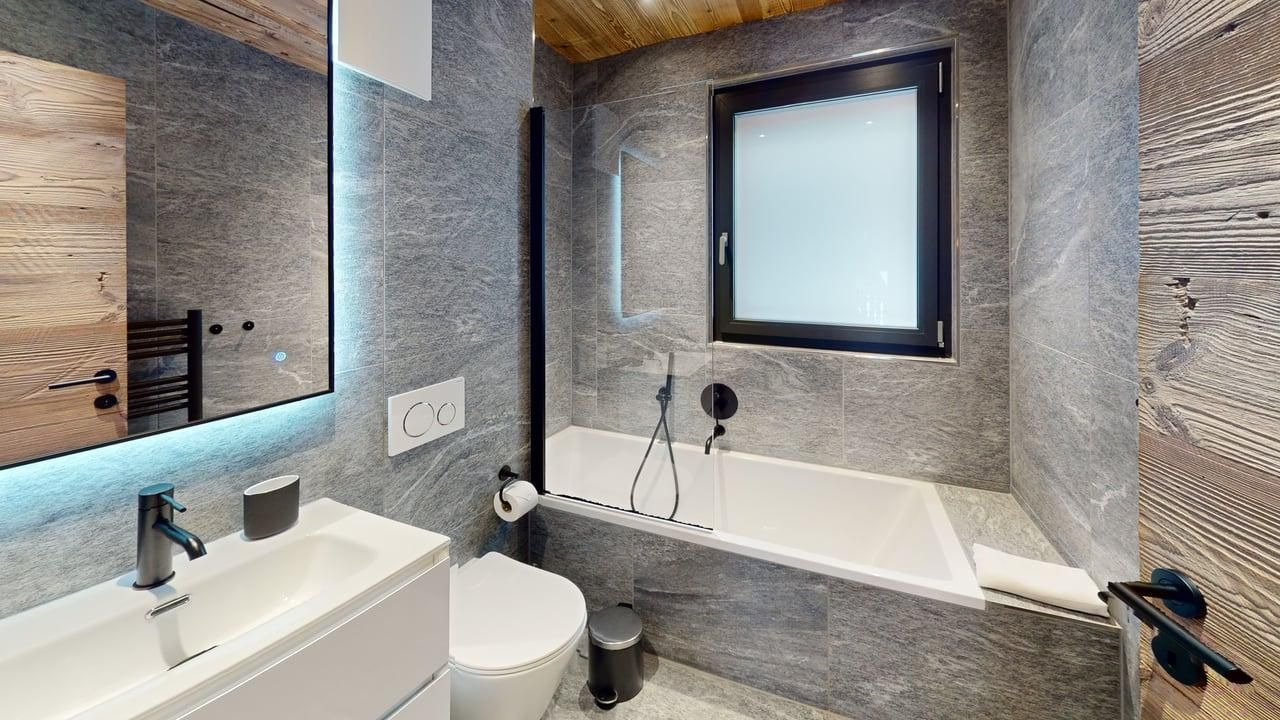 A modern bathroom with gray stone tiles, a white vanity with a rectangular sink, a toilet, and a bathtub with a window.