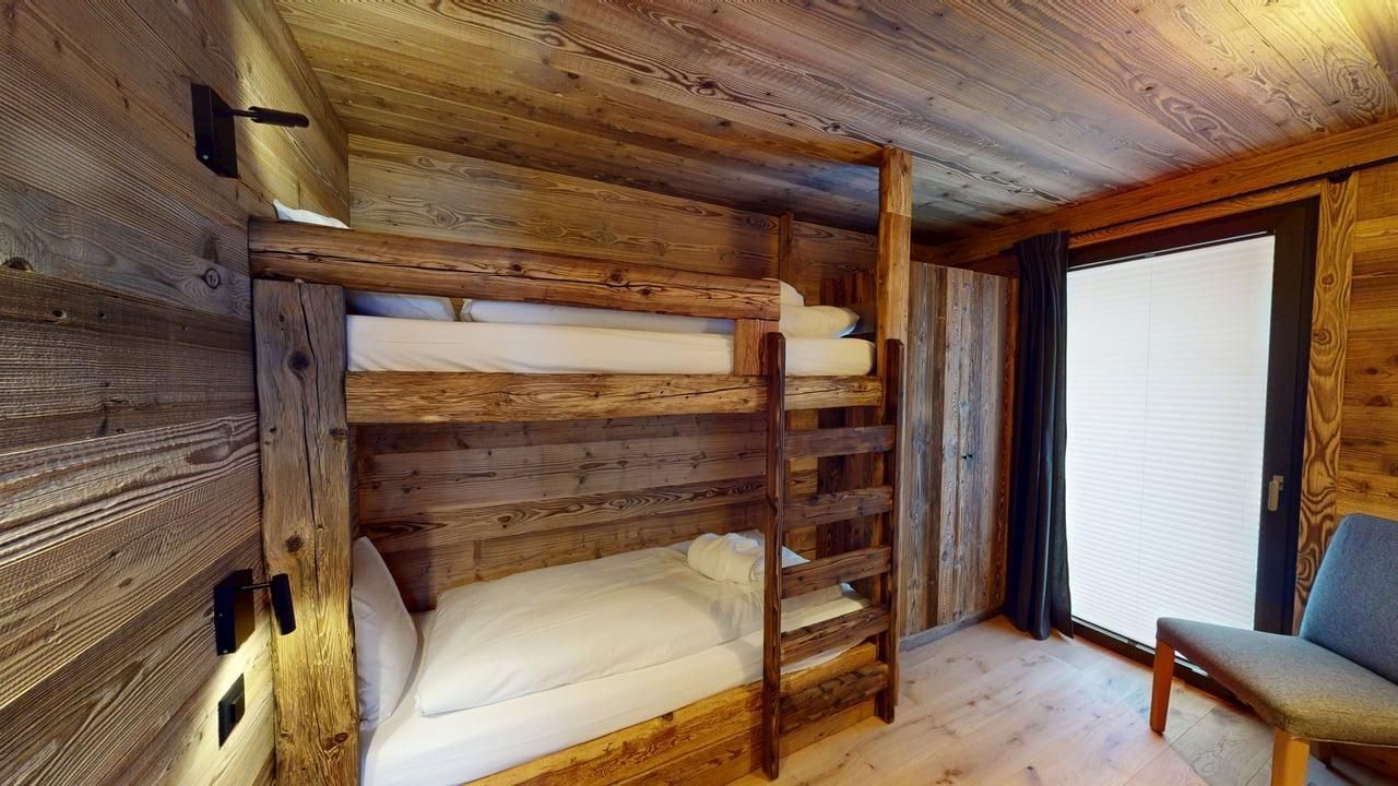 A rustic wooden bunk bed in a cabin-style bedroom with a white window blind and a gray armchair.