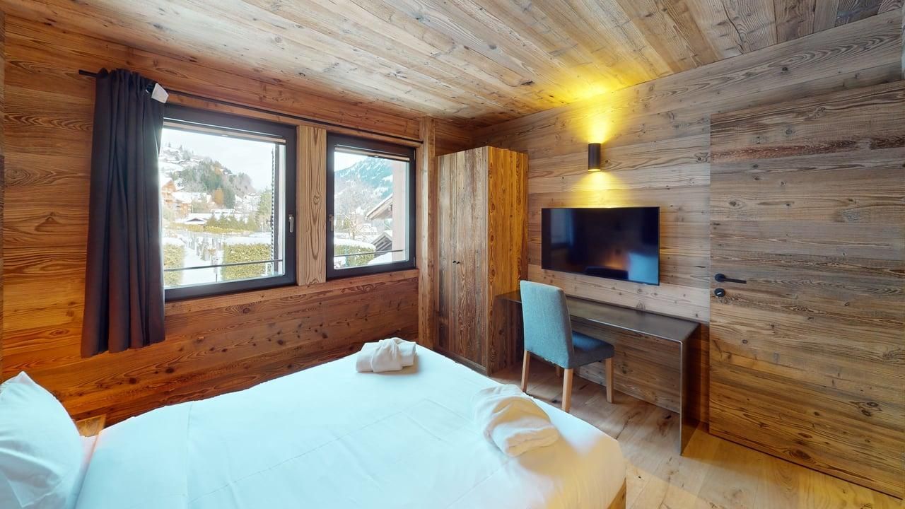 A rustic hotel bedroom with wooden walls, a bed, a window, a wooden desk with a chair, and a wall-mounted TV.