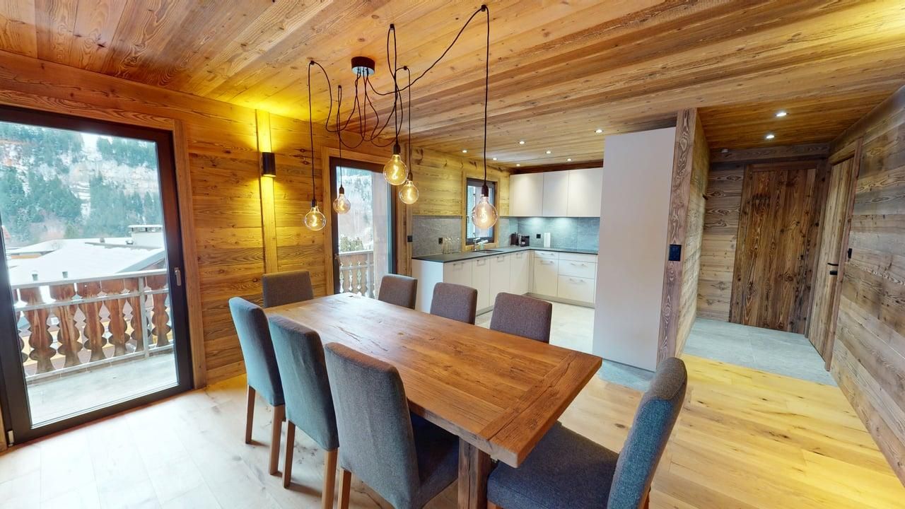 A modern wooden dining room features a large table with six gray chairs, a unique pendant light, and an open-plan kitchen.