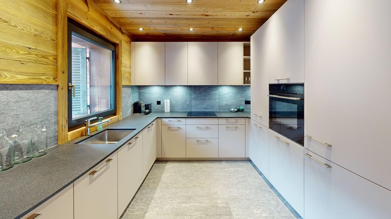 A modern kitchen with light-colored cabinets, granite countertops, stone backsplash, and a wooden ceiling.