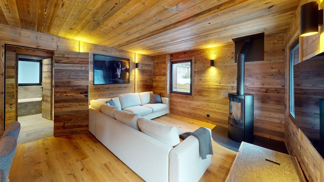 A cozy living room with wooden walls, a light-colored sofa, a fireplace, and an adjacent bathroom.