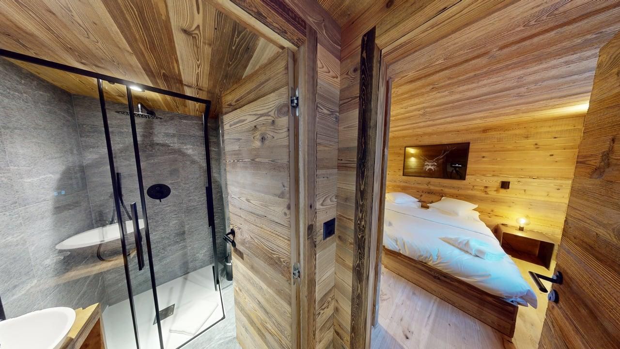 Vue depuis une salle de bains rustique avec douche vitrée donnant sur une chambre confortable aux murs lambrissés.