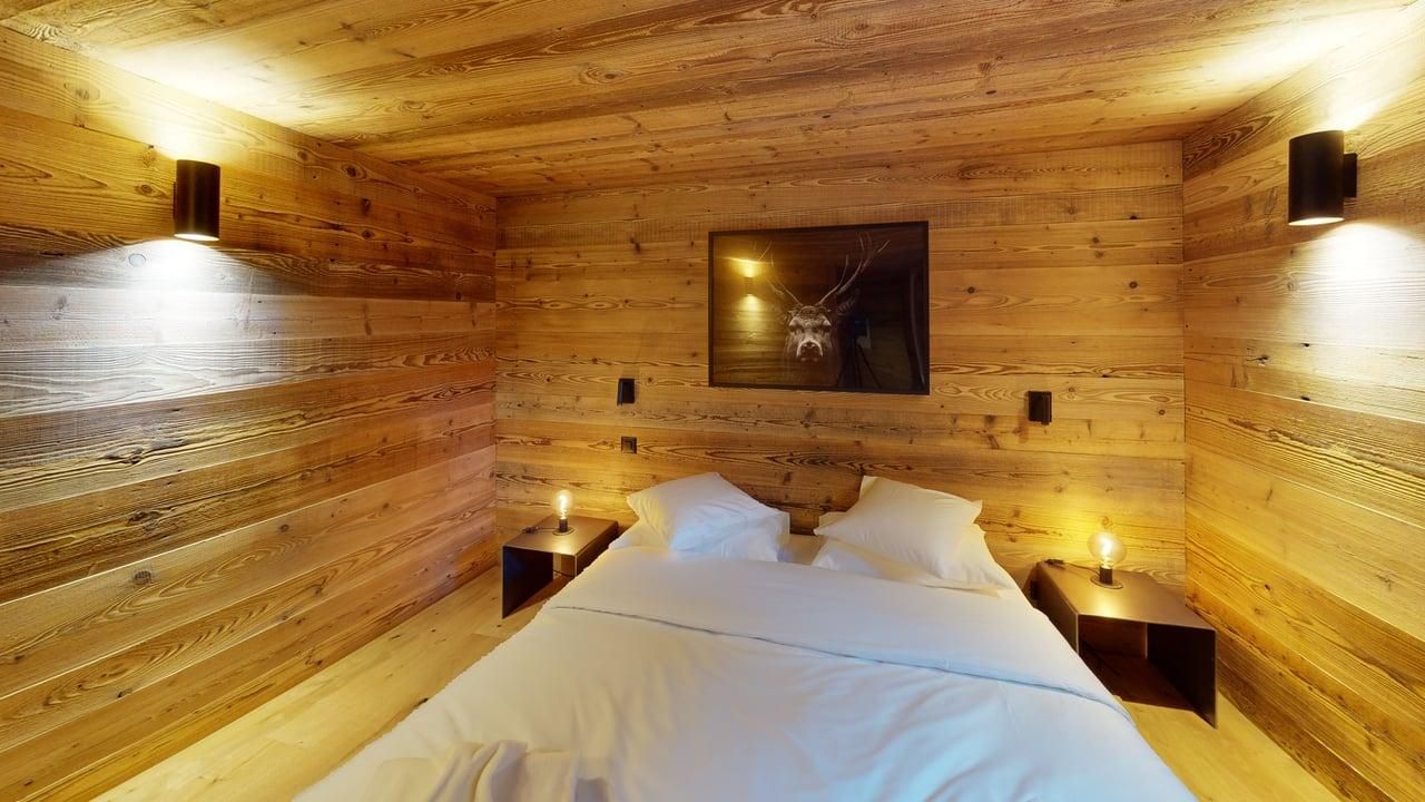 Une chambre avec des murs lambrissés, un lit recouvert de draps blancs, deux petites tables de chevet et deux appliques murales.