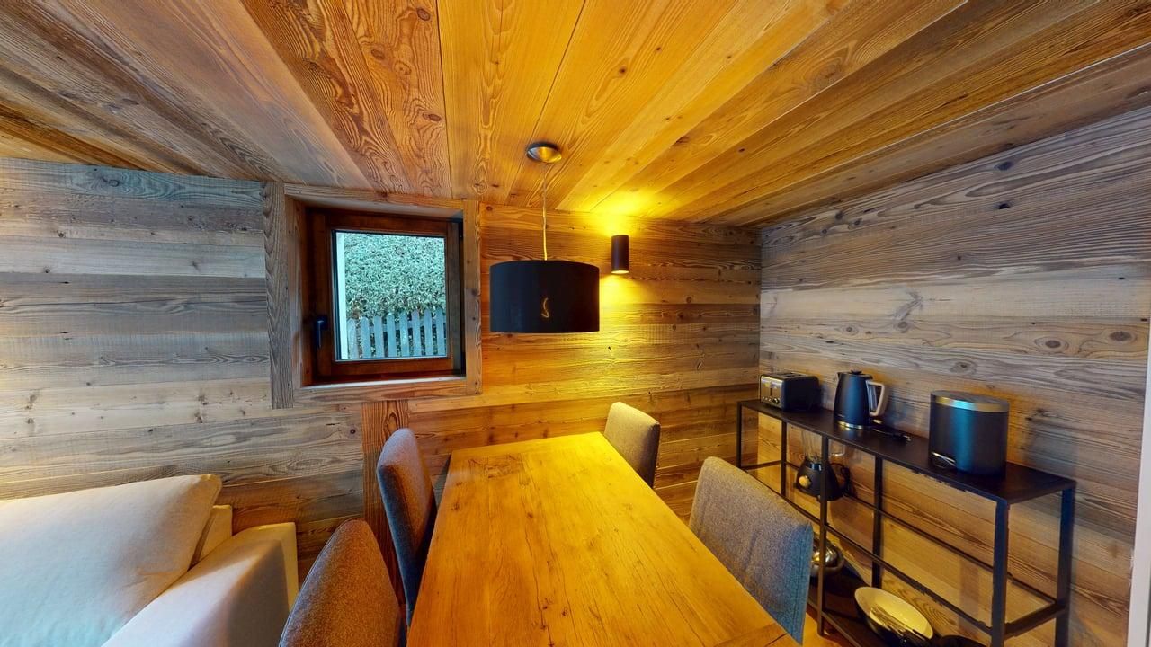 Un coin repas chaleureux, lambrissé, avec une table en bois, des chaises grises, une suspension noire et une petite fenêtre.