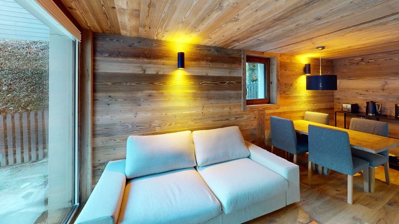 Un intérieur lumineux, tout en bois, comprend un canapé blanc, une table à manger avec des chaises grises et un éclairage mural chaleureux.