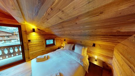 Une chambre mansardée confortable en bois, avec un lit, du linge de lit blanc, un éclairage d'ambiance chaleureux et une porte-fenêtre donnant sur un balcon avec vue sur la neige.