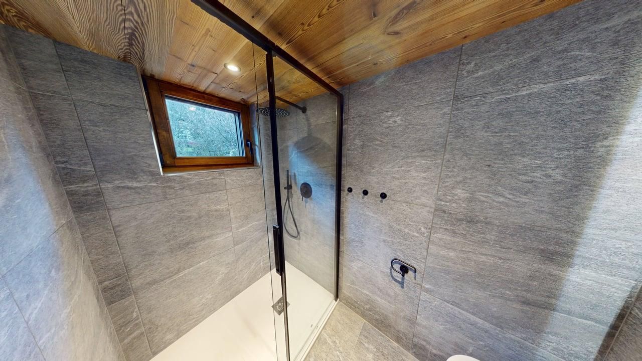 A modern bathroom with a glass-enclosed shower, gray tile walls, and a wooden ceiling with a small window.