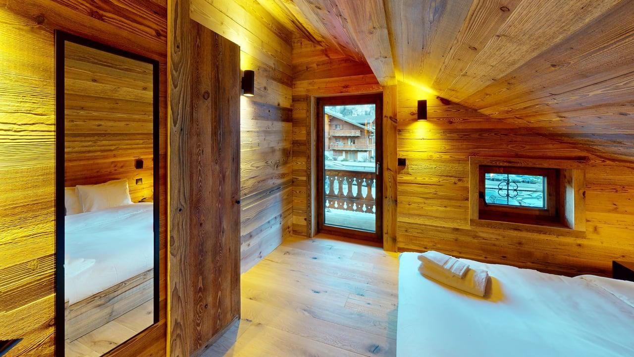 A rustic bedroom with wood-paneled walls, a large mirror, two white beds, and a glass door leading to a balcony.